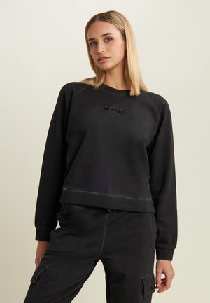 Giovane donna con capelli biondi lisci, che indossa una felpa nera e pantaloni cargo neri, su uno sfondo chiaro e semplice.