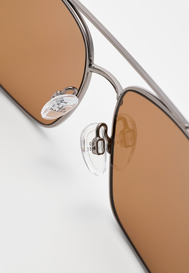 Eine Sonnenbrille mit braunen Gläsern, einem Metallrahmen und klaren Nasenpolstern. Der Rahmen verfügt über ein schlankes, abgerundetes Design und einen minimalistischen Stil.