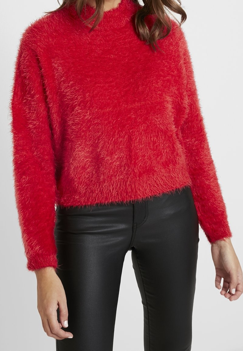 Femme portant un pull rouge duveteux à manches longues et un pantalon en cuir noir, debout devant un fond clair uni.