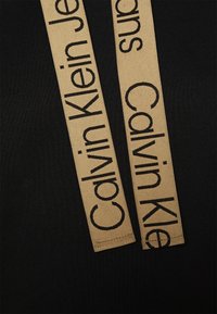 Czarna tkanina z dwoma beżowymi paskami z napisem "Calvin Klein Jeans" pogrubionymi czarnymi literami. Paski są gładkie i przylegają płasko do materiału.