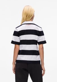 Polo s krátkým rukávem s černobílými vodorovnými pruhy, s pevným límcem a červeným logem na zadní straně krku. Jemný textil.