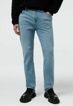 Mężczyzna w jasnoniebieskich prostych dżinsach, czarnym pasku, czarnych masywnych butach sznurowanych i czarnej bluzce z długim rękawem, z rękami w kieszeniach.