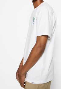 Mies, joka on pukeutunut valkoiseen lyhythihaiseen t-paitaan, jossa on pieni graafinen kuva rinnassa, ja beigeihin shortseihin, seisoo yksinkertaista valkoista taustaa vasten.