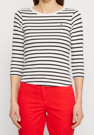 Vrouw draagt een wit-zwart gestreept shirt met lange mouwen en een klein logo op de borst, gecombineerd met felrode broek.
