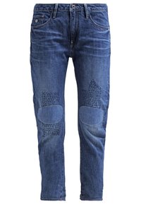 Jeans de hombre en denim azul con detalles de patchwork y tela texturizada en las áreas de las rodillas, bolsillos frontales, trabillas y cierre de botón.