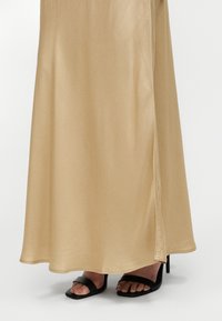 Jupe beige longueur cheville avec fente latérale portée avec des sandales à talons hauts noires à bout ouvert sur fond blanc.