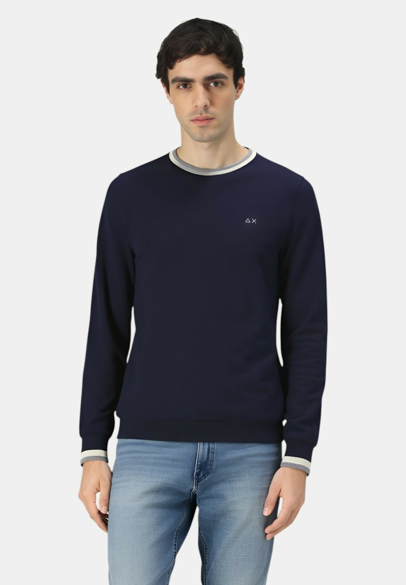 Uomo che indossa un felpa a maniche lunghe blu navy con colletto e polsini a strisce bianche, abbinata a jeans azzurri, in piedi con il volto rivolto in avanti.