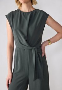 Jumpsuit verde scuro a wrap con maniche corte, dettaglio con nodo frontale e texture liscia. Presenta una vestibilità rilassata e una silhouette fluida.