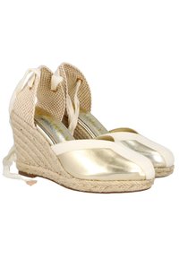 Alpargatas de cuña con puntera metálica dorada, parte superior de tela cremosa, suela de yute trenzado y cintas en los tobillos. Diseño texturizado con talón abierto.