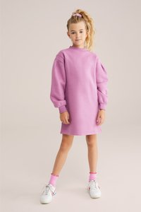 Roza dukserica haljina s dugim rukavima i rebrastim manžetama. Sadrži diskretni izvezeni logotip na prsima. Uparena s bijelim tenisicama i ružičastim čarapama.