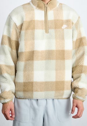 Homme portant un pull en polaire beige et blanc à carreaux avec une fermeture éclair au quart et un pantalon de survêtement gris clair.