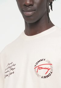 Μπεζ βαμβακερό T-shirt με στρογγυλή λαιμόκοψη, διαθέτει τυπωμένο κείμενο και λογότυπα σε καφέ και κόκκινο. Κοντά μανίκια, άνετη εφαρμογή, λεία υφή.