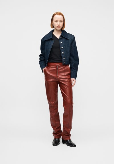 Γυναίκα που φοράει κοντό σκούρο τζιν μπουφάν, μαύρο τοπ, καφέ δερμάτινο παντελόνι με φερμουάρ μπροστά και μαύρα μπότες με τακούνι, στέκεται με τα χέρια στις τσέπες.