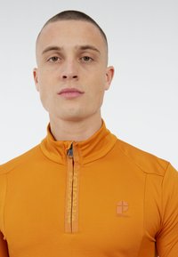 Jaunas vyras su trumpais plaukais, vilkintis aukštą apykaklę apelsinų spalvos užtrauktuku marškinį su užrašu ir logotipu „PROTEST“ krūtinėje.