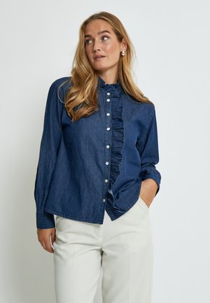 Žena s dugom plavom kosom, odjevena u tamnoplavu košulju s volanima i bijele hlače, pozira s jednom rukom u džepu.