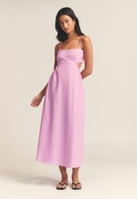 Abito maxi a taglio con tessuto rosa pastello, spalline sottili, scollatura quadrata, ritagli laterali e gonna fluida. Abbinato a sandali neri.