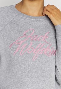 Felpa grigia con il logo ricamato rosa di "Jack Wolfskin" sul petto, con una texture morbida e maniche raglan.