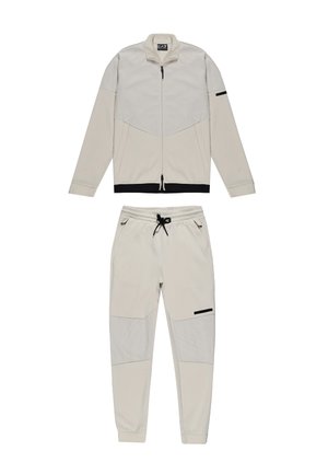 Survêtement de couleur claire composé d'une veste zippée avec un design en maille et d'un pantalon assorti. Les deux incorporent des accents noirs et des poignets élastiques.
