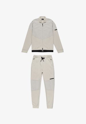 Survêtement de couleur claire composé d'une veste zippée avec un design en maille et d'un pantalon assorti. Les deux incorporent des accents noirs et des poignets élastiques.