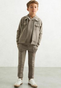Hellbrauner Kapuzenpullover mit Fronttaschen, kombiniert mit passenden Jogginghosen. Verfügt über gerippte Bündchen und eine weiche Textur. Abgerundet mit weißen Sneakers.