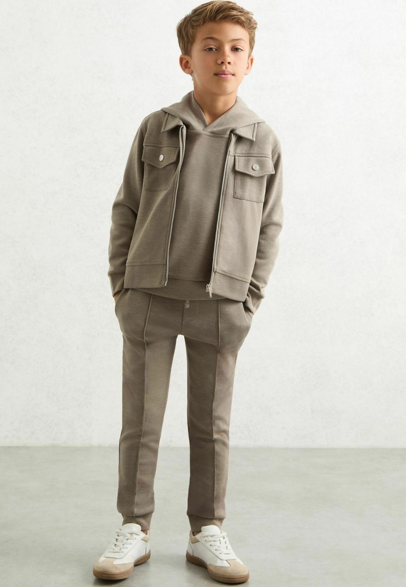 Hellbrauner Kapuzenpullover mit Fronttaschen, kombiniert mit passenden Jogginghosen. Verfügt über gerippte Bündchen und eine weiche Textur. Abgerundet mit weißen Sneakers.
