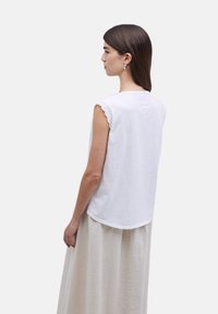 Camisa sin mangas de algodón blanca con bordes de mangas en forma de onda, con cuello redondo y dobladillo curvado, combinada con una falda beige.