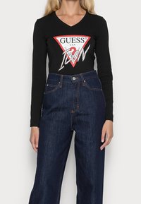 Černé tričko s dlouhým rukávem a červeným trojúhelníkovým logem, na kterém je bíle napsáno "GUESS" a "ICON", v kombinaci s tmavě modrými džíny s vysokým pasem.