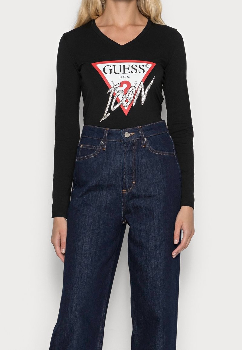 Černé tričko s dlouhým rukávem a červeným trojúhelníkovým logem, na kterém je bíle napsáno "GUESS" a "ICON", v kombinaci s tmavě modrými džíny s vysokým pasem.