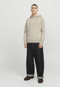 Beige hoodie with a front logo, paired with loose-fitting dark denim jeans, rolled cuffs, and brown shoes. Simple and casual design.