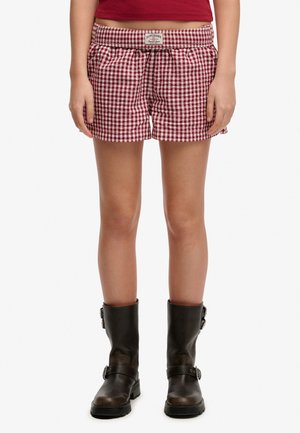 Persoon draagt rood-wit geruit korte broekje, rood cropped topje en zwarte laarzen tot halverwege de kuit met gespen, staand op witte achtergrond.