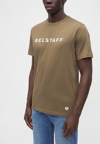 Άνδρας που φοράει ελιά πράσινη μπλούζα Belstaff και μπλε τζιν, στέκεται μπροστά σε απλό φωτεινό φόντο.