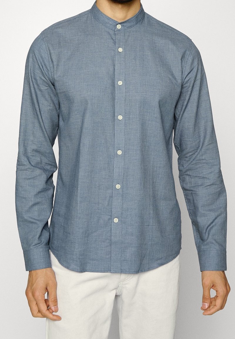 Camicia a maniche lunghe con bottoni in tessuto blu chiaro con una trama texture, dotata di colletto alla mandarina e bottoni bianchi.