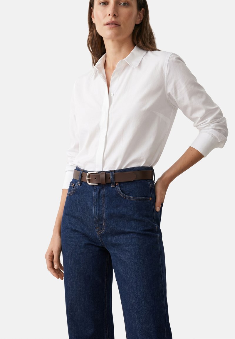 Frau trägt ein weißes Langarmshirt, das in dunkelblaue Jeans mit braunem Gürtel gesteckt ist, eine Hand in der Tasche, vor schlichtem Hintergrund.