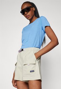 Lys blå kortærmet skjorte og beige cargo shorts med bæltested. Bemærkelsesværdige funktioner inkluderer to sidelommer og et logo-badge på shortsene.