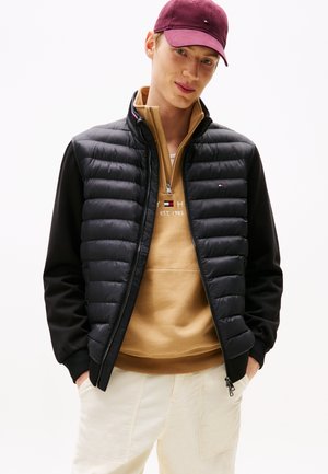 Ung mand iført en vinrød kasket, sort quiltet jakke, tan hættetrøje og hvide bukser, poserer med hænderne i lommerne mod en hvid baggrund.