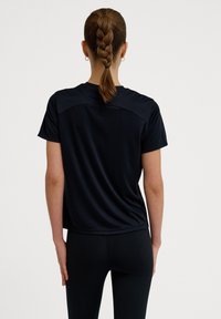 Sort atletisk t-shirt med korte ærmer, glat tekstur, med en let glans og detaljer ved skuldersømmen, kombineret med sorte tights.