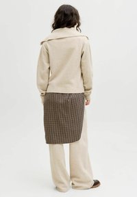 Persona che indossa un maglione beige e pantaloni con un pannello a pieghe a scacchi neri e beige sul retro, in piedi su uno sfondo bianco uniforme.