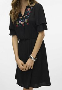 Svart kortärmad klänning med V-ringning, blomsterbroderi i rött, blått och grönt, med gathered midja och flödande kjol. Silverarmband.