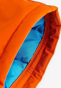Tela exterior naranja con una textura suave; forro interior en azul brillante, acolchado para aislamiento; detalle de costura notable alrededor del borde.
