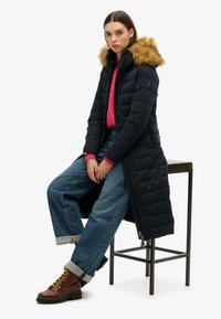 Marineblauwe lange pufferjas met een afneembare nepbont capuchon, gedragen over een roze trui, gecombineerd met losse blauwe jeans en bruine veterschoenen.