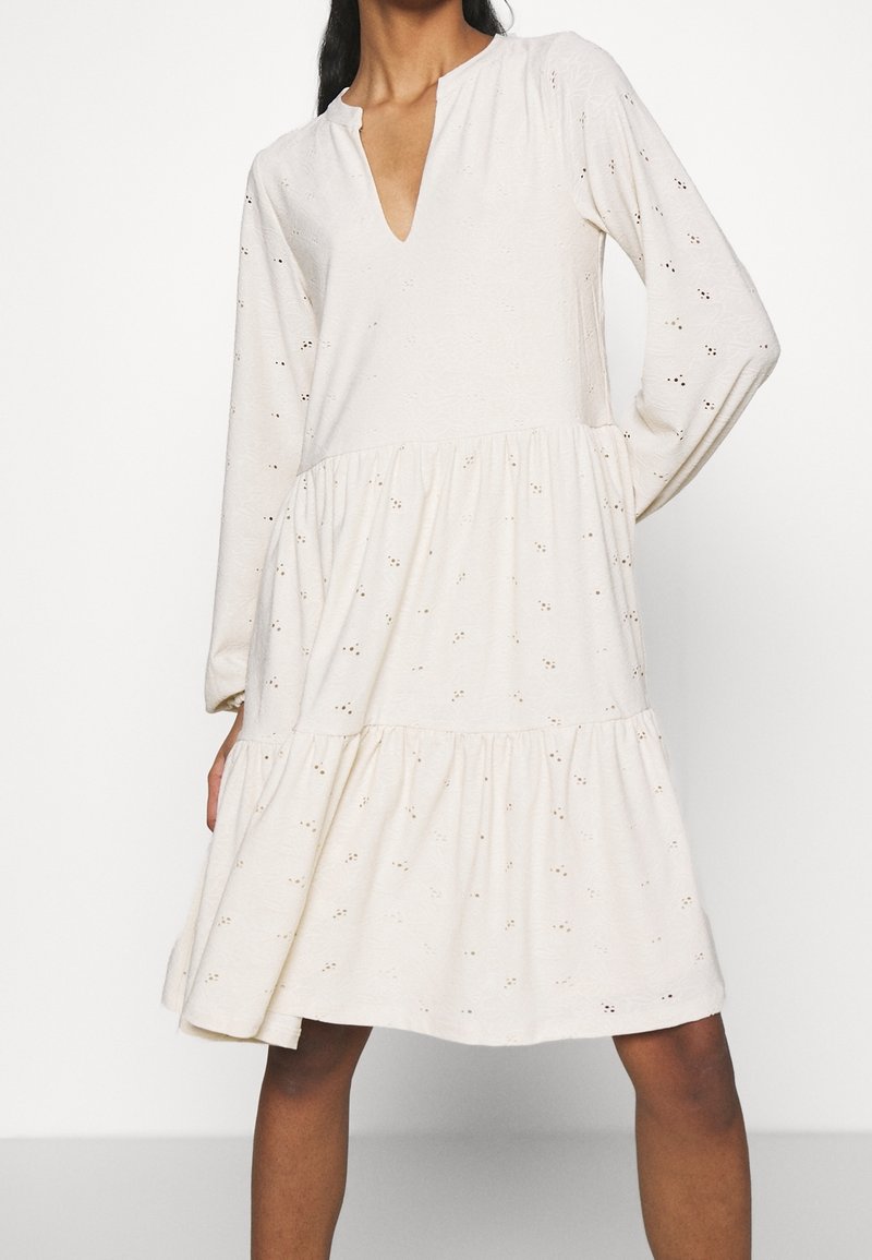 Femme portant une robe ample de couleur crème à manches longues, avec des détails en œillets et un col en V, debout devant un fond blanc.