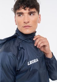 Giacca a vento blu navy con colletto alto, chiusura con zip, polsini elasticizzati e logo bianco "LEGEA" sul petto. Tessuto liscio.