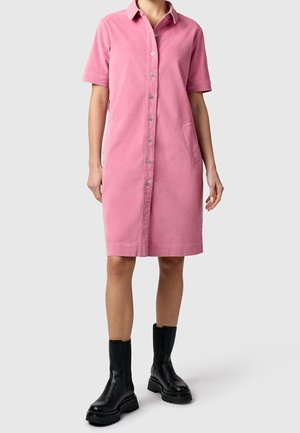 Personne portant une robe rose boutonnée à manches courtes avec des poches, associée à des bottes noires épaisses montant jusqu'à la moitié du mollet sur un fond clair.