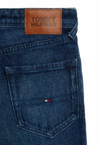 Jeans de denim azul oscuro con una etiqueta de cuero grabada con "TOMMY HILFIGER" y un pequeño acento rojo, blanco y azul cerca del bolsillo trasero.