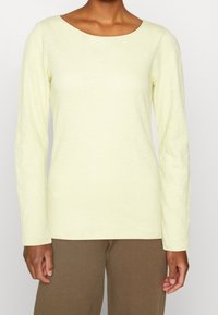 Helles gelbes Langarmshirt mit Rundhalsausschnitt, aus einem weichen, strukturierten Stoff. Getragen mit braunen Hosen, der Arm ist links sichtbar.