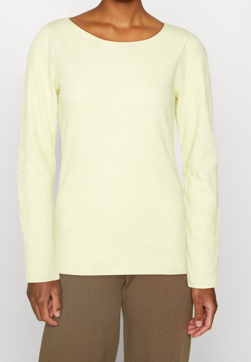 Helles gelbes Langarmshirt mit Rundhalsausschnitt, aus einem weichen, strukturierten Stoff. Getragen mit braunen Hosen, der Arm ist links sichtbar.