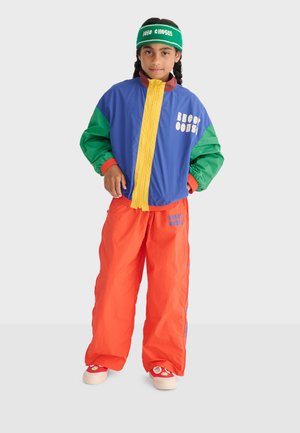 Niño con cabello trenzado vistiendo chaqueta rompevientos colorida, pantalones naranjas, zapatillas rojas y una diadema verde con texto, de pie contra un fondo blanco.
