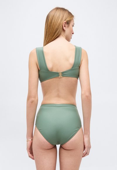 Γυναίκα με μακριά ξανθά μαλλιά που φοράει ένα πράσινο μαγιό με ψηλόμεσο κάτω μέρος και ταιριαστό τοπ με χρυσό κούμπωμα, στέκεται με την πλάτη γυρισμένη πάνω σε λευκό φόντο.