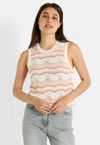 Top de punto sin mangas en blanco con patrones de olas naranjas. Presenta un cuello redondo y una textura de tejido abierto. Combinado con unos jeans de color azul claro.