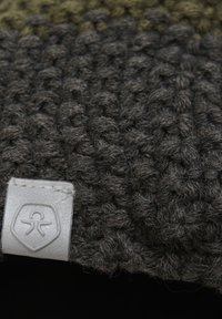 Tjockstickad halsduk i mörkgrå ull med en sektion i olivgrönt, prydd med en silverfärgad märkesetikett sydd på kanten. Texturerad yta synlig.